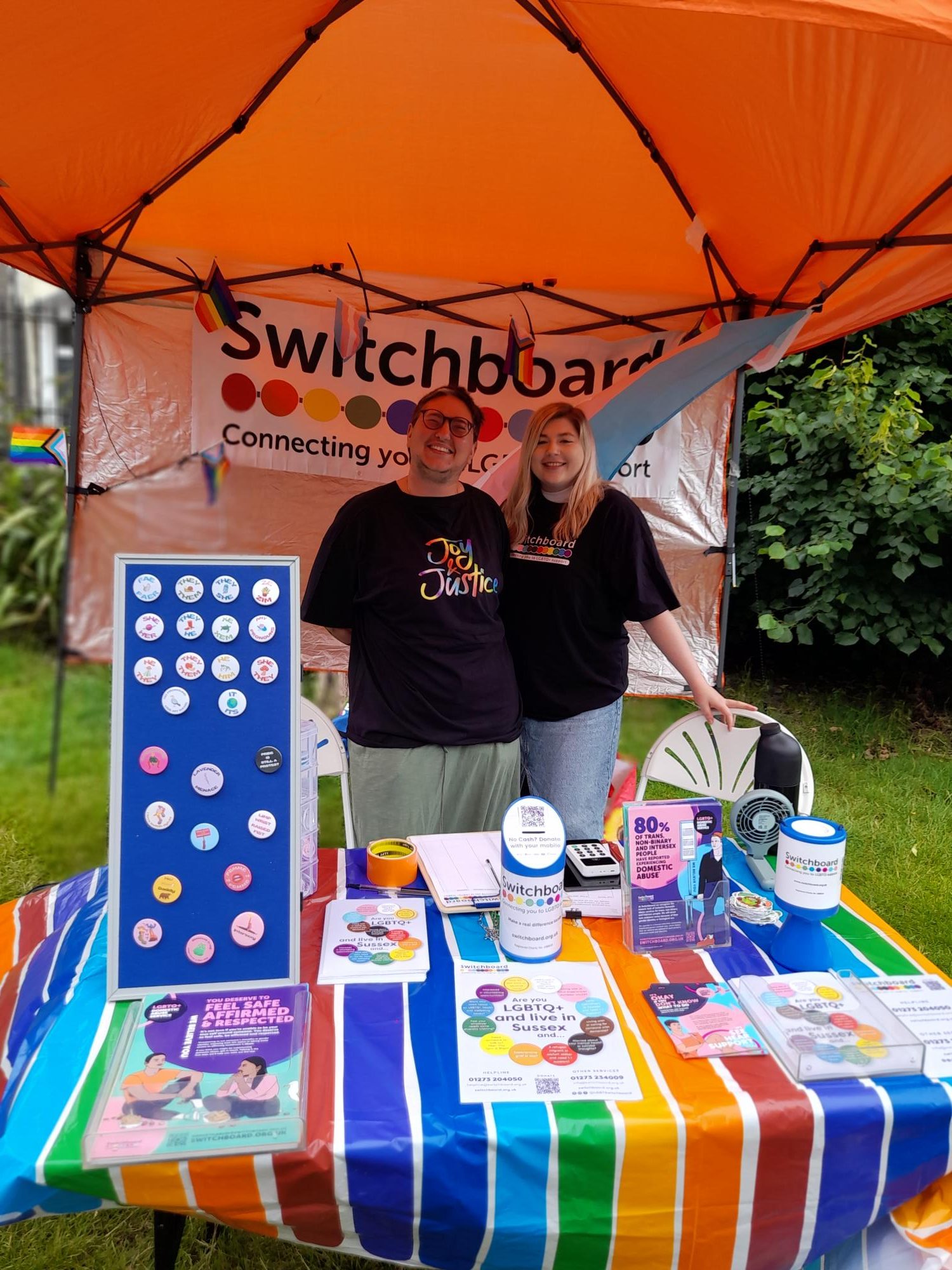 A photo of Raf (they/them) and Ciara (she/her) at Switchboard's stall for Trans Pride. They wear Switchboard's t-shirts, Raf's says "Joy & Justice" and Ciara's has Switchboard's logo on. In front of them are Switchboard's pronoun badges and leaflets advertising our services.