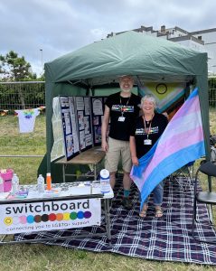 A photo of Chris (he/him) and Jane (she/her) standing together at Switchboard's stall for Eastbourne Pride. They are wearing black t-shirts with Switchboard's logo on. Jane is holding a large Trans Pride Flag. In front of the stall, there is a table with leaflets, pronoun badges and a banner advertising Brighton & Hove LGBT Switchboard.