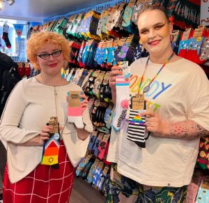 Brighton & Hove LGBT Switchboard team members Emily and Astrid in the Brighton Soctopus store. They are smiling and holding the socks from Soctopus's Pride sock range, which include Progress Flag socks, Trans Pride flag socks, and Ally Flag socks.