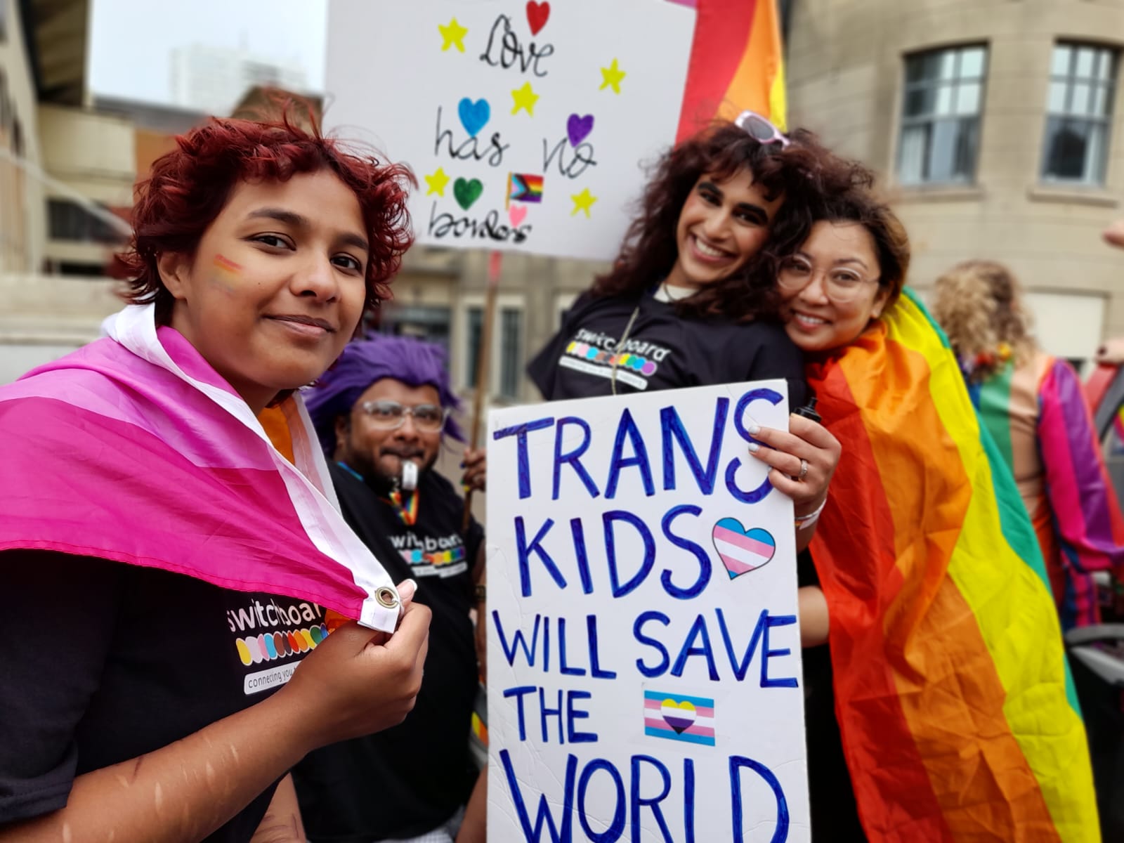 4 Switchboard volunteers and guests hold signs that say "Love Has No Borders" and "Trans Kids Will Save The World". They wear Switchboard t-shirts, and two wear flags on their backs: the Lesbian Pride Flag and the Pride flag.