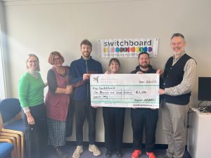 A photograph of Shutterly Fabulous and Brighton & Hove LGBT Switchboard employees holding a cheque for £1,700 from Shutterly Fabulous for Brighton & Hove LGBT Switchboard. Switchboard's logo is behind them.