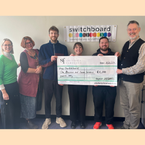 A photograph of Shutterly Fabulous and Brighton & Hove LGBT Switchboard employees holding a cheque for £1,700 from Shutterly Fabulous for Brighton & Hove LGBT Switchboard. Switchboard's logo is behind them.